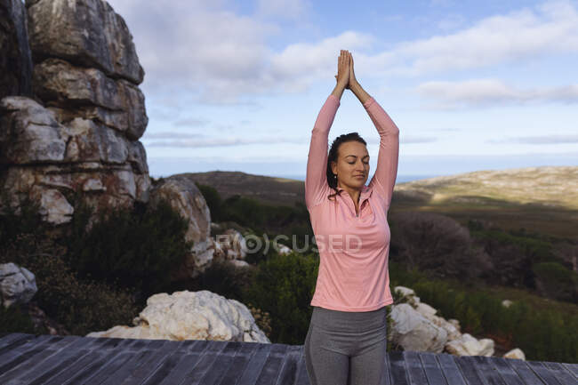 Mujer caucásica sonriente practicando yoga de pie con los brazos levantados en el entorno rural de montaña. vida sana, fuera de la red y cerca de la naturaleza. - foto de stock