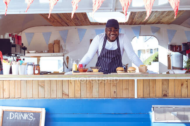 Portrait of smiling african american man in food truck with hamburgers on worktop. independent business and street food service concept. — Stock Photo