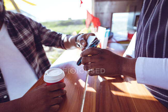 Midsection of african american man in food truck taking smartphone payment holding terminal. independent business and street food service concept. — Stock Photo