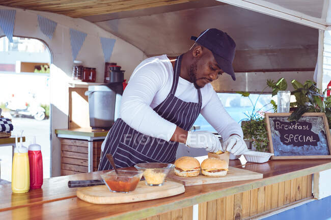 African american man in food truck preparing order with hamburgers on worktop. independent business and street food service concept. — Stock Photo