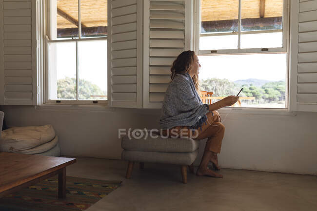 Счастливая белая женщина в наушниках с помощью смартфона сидит у окна в коттеджной гостиной. простая жизнь в глуши сельских домов. — стоковое фото