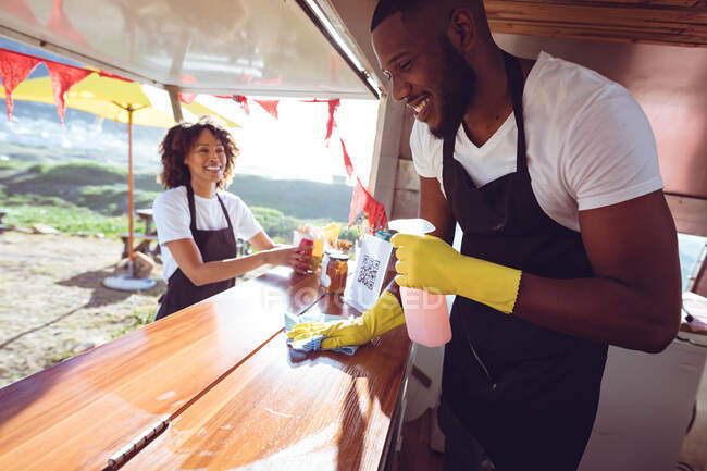 Smiling diverse couple behind cleaning counter in food truck. independent business and street food service concept. — Stock Photo