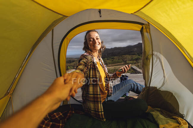 Smiling caucasian woman camping, sitting outside on mountainside deck holding hand of friend in tent. healthy living, off the grid and close to nature. — Stock Photo