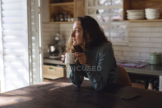 Mujer caucásica feliz de pie en la cocina de la cabaña apoyada en el mostrador sosteniendo el café mirando hacia otro lado. simple vivir en una casa rural fuera de la red. - foto de stock