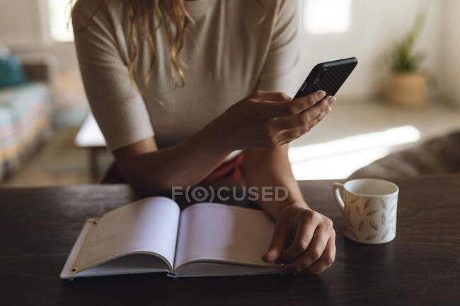 Sección media de la mujer sentada en el escritorio con el libro y el café con el teléfono inteligente. trabajar en casa aislado durante el bloqueo de cuarentena. - foto de stock