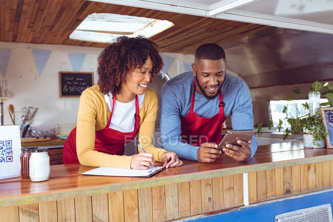 Sonriente pareja diversa detrás del mostrador utilizando la tableta en camión de alimentos. concepto de empresa independiente y servicio de comida callejera. - foto de stock