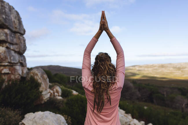 Vista trasera de una mujer caucásica practicando yoga sentada con los brazos levantados en un entorno rural de montaña. vida sana, fuera de la red y cerca de la naturaleza. - foto de stock