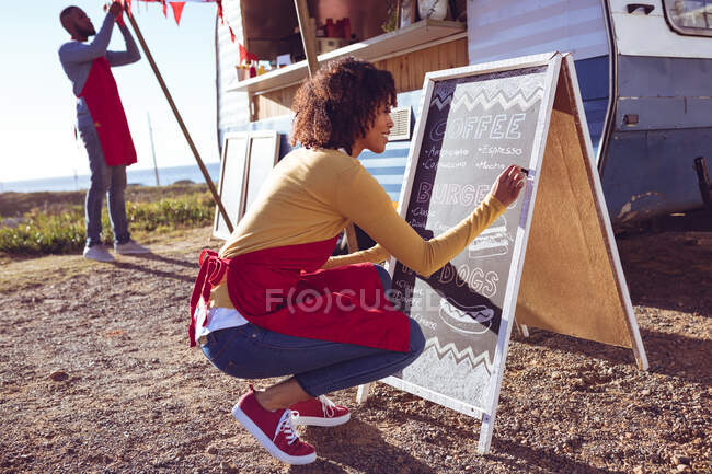 Diverse couple opening and preparing food truck by seaside on sunny day, woman writing on menu board. independent business and street food service concept. — Stock Photo