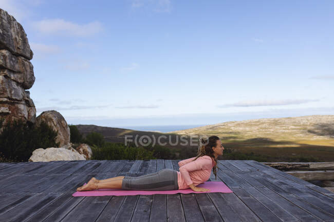 Mulher caucasiana feliz praticando ioga deitado no convés que se estende no cenário de montanha rural. vida saudável, fora da grade e perto da natureza. — Fotografia de Stock