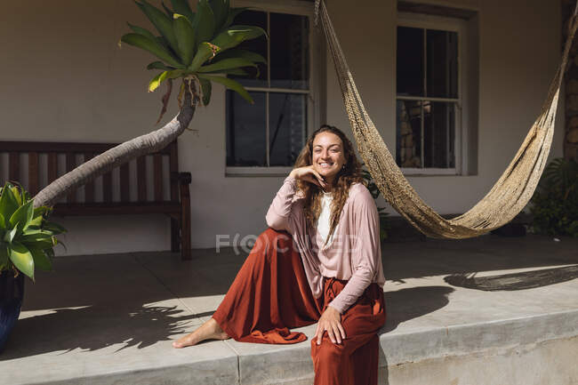 Femme caucasienne souriante assise relaxante sur la terrasse ensoleillée du chalet. mode de vie sain, proche de la nature dans la maison rurale hors réseau. — Photo de stock