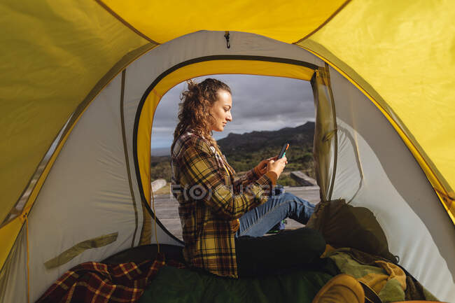 Mulher caucasiana acampar, sentado fora da tenda no deck de montanha usando smartphone. vida saudável, fora da grade e perto da natureza. — Fotografia de Stock