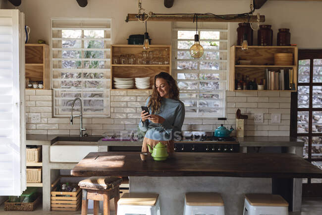 Felice donna caucasica in piedi in cucina cottage utilizzando smartphone e sorridente. semplice vivere in una casa rurale fuori dalla griglia. — Foto stock