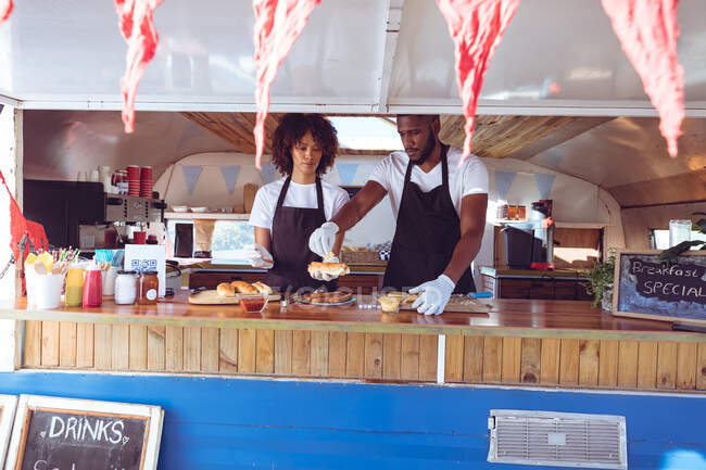 Diverse couple preparing hot dogs behind counter in food truck. independent business and street food service concept. — Stock Photo