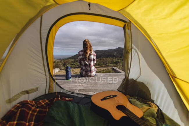 Vista trasera de la mujer caucásica acampando, sentada fuera de la tienda en la cubierta de la montaña admirando la vista. vida sana, fuera de la red y cerca de la naturaleza. - foto de stock