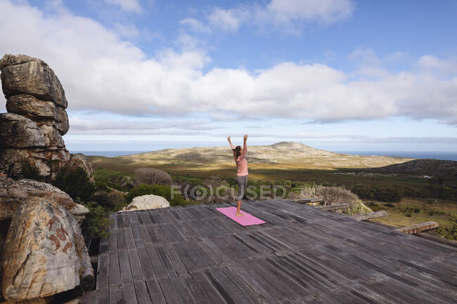 Mujer caucásica practicando yoga de pie sobre una pierna estirándose en un entorno rural de montaña. vida sana, fuera de la red y cerca de la naturaleza. - foto de stock
