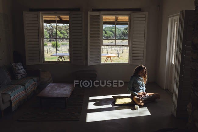 Happy caucasian woman sitting on floor using laptop in sunny cottage living room. simple living in an off the grid rural home. — Stock Photo