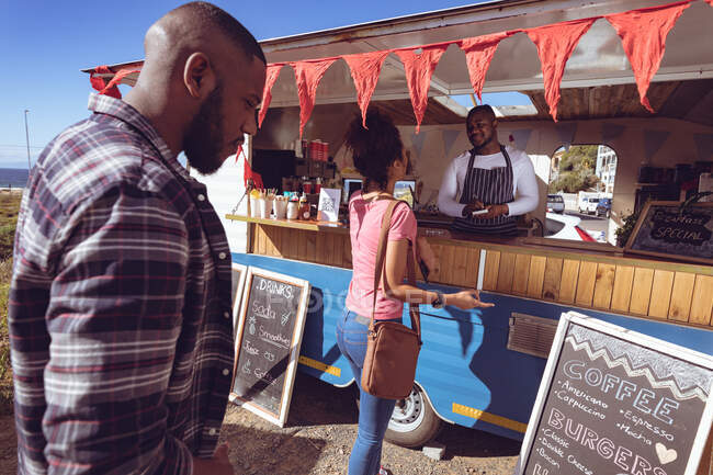 Улыбающийся африканский американец в фургоне с едой принимает заказ от женщины-клиента. независимая концепция обслуживания бизнеса и уличной еды. — стоковое фото