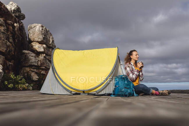 Mulher caucasiana feliz acampar, sentado segurando café fora da tenda no deck de montanha. vida saudável, fora da grade e perto da natureza. — Fotografia de Stock