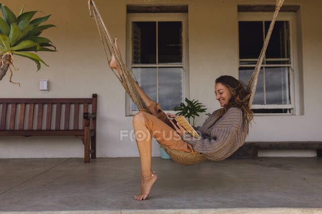 Felice donna caucasica seduta in amaca sulla terrazza cottage, leggere libro e sorridere. vita sana, vicino alla natura fuori dalla griglia casa rurale. — Foto stock