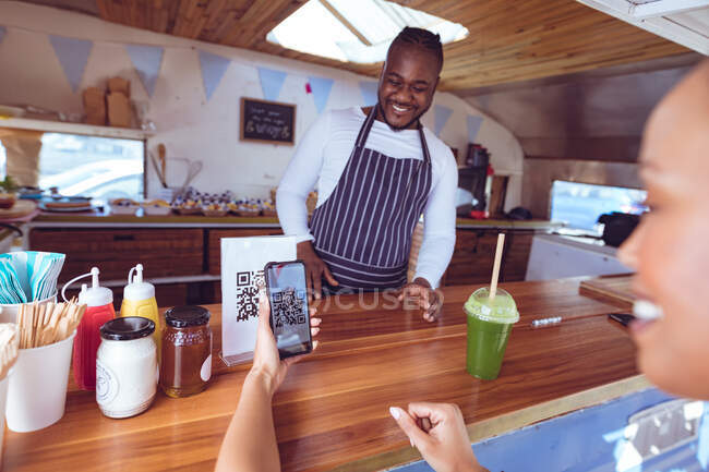 Smiling african american man in food truck with female customer reading qr code with smartphone. independent business and street food service concept. — Stock Photo