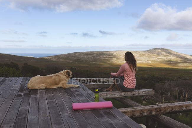 Mulher caucasiana pensativo sentado no convés com cão de estimação admirando a vista no cenário montanhoso rural. vida saudável, fora da grade e perto da natureza. — Fotografia de Stock