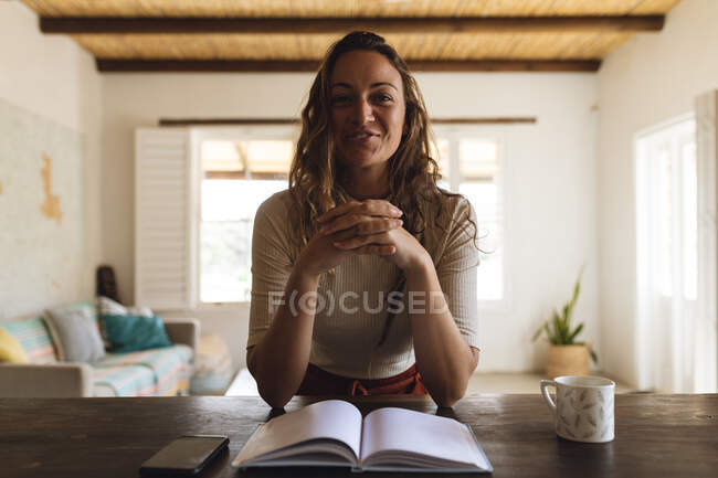 Mujer caucásica sonriente sentada en el escritorio con libro y café haciendo videollamada. trabajar en casa aislado durante el bloqueo de cuarentena. - foto de stock
