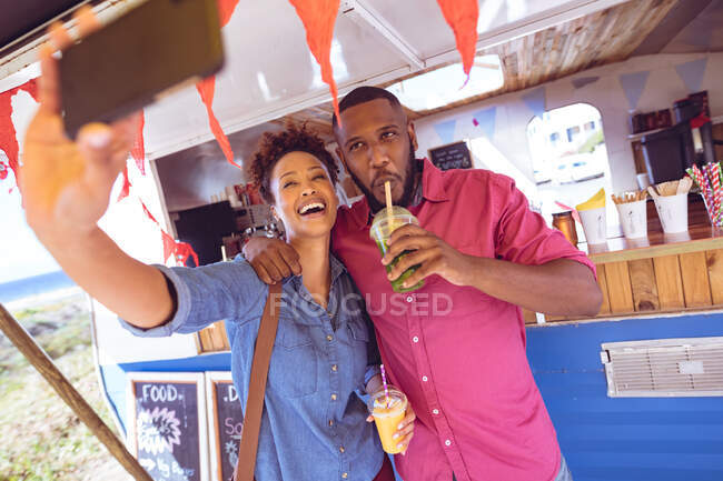 Smiling diverse couple taking selfie with smartphone and having drink by food truck at seaside. independent business and street food service concept. — Stock Photo