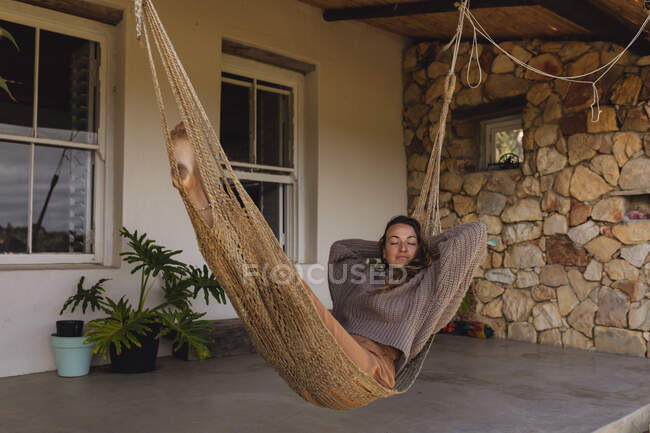 Heureuse femme caucasienne allongée dans un hamac sur la terrasse du chalet, relaxante avec les yeux fermés. mode de vie sain, proche de la nature dans la maison rurale hors réseau. — Photo de stock