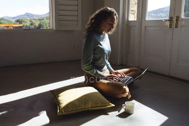 Mulher caucasiana feliz sentado no chão usando laptop na sala de estar ensolarada casa de campo. simples viver em uma casa rural fora da grade. — Fotografia de Stock