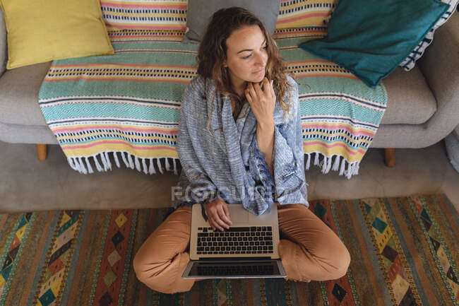 Femme caucasienne réfléchie avec couverture assise sur le sol en utilisant un ordinateur portable dans le salon ensoleillé du chalet. vivre simplement dans une maison rurale hors réseau. — Photo de stock