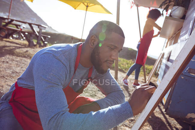 Smiling african american man writing on menu board next to food truck on sunny day. independent business and street food service concept. — Stock Photo