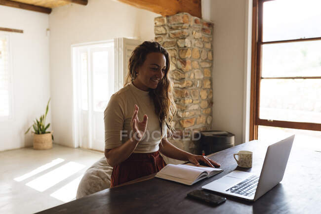 Белая женщина сидит за столом с книгой, используя ноутбук, делает видеозвонок и машет рукой. работа на дому в изоляции во время карантинной изоляции. — стоковое фото