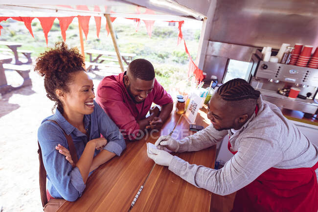Homme afro-américain souriant dans un camion de nourriture bavardant avec des clients masculins et féminins. entreprise indépendante et concept de service de restauration de rue. — Photo de stock