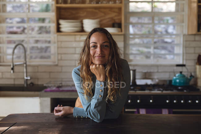 Retrato de mujer caucásica feliz de pie en la cocina de la cabaña apoyada en el mostrador y sonriendo. simple vivir en una casa rural fuera de la red. - foto de stock