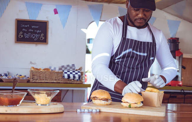 Hombre afroamericano en camión de comida preparando orden con hamburguesas en la encimera. concepto de empresa independiente y servicio de comida callejera. - foto de stock