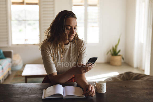 Кавказка сидит за столом с книгой и кофе, используя смартфон. работа на дому в изоляции во время карантинной изоляции. — стоковое фото