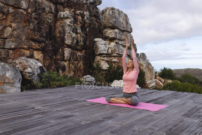 Feliz mujer caucásica practicando yoga sentada en cubierta estirándose en un entorno rural de montaña. vida sana, fuera de la red y cerca de la naturaleza. - foto de stock