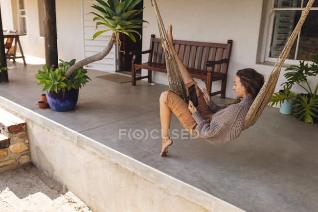 Mujer caucásica feliz relajarse en hamaca en la terraza casa de campo, utilizando la tableta. vida sana, cerca de la naturaleza en el hogar rural fuera de la red. - foto de stock