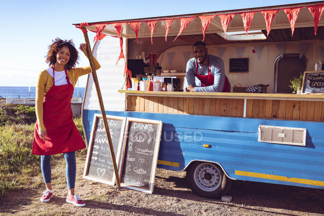 Portrait of smiling diverse couple in food truck by seaside. independent business and street food service concept. — Stock Photo