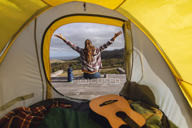 Вид сзади на кемпинг кавказской женщины, сидящей снаружи палатки на склоне горы с вытянутыми руками. здоровый образ жизни, вне сети и близко к природе. — стоковое фото