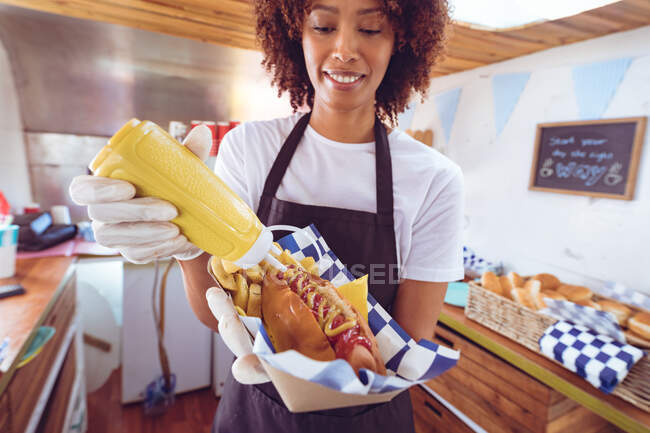 Smiling mixed race woman pouring mustard on hot dog behind counter in food truck. independent business and street food service concept. — Stock Photo