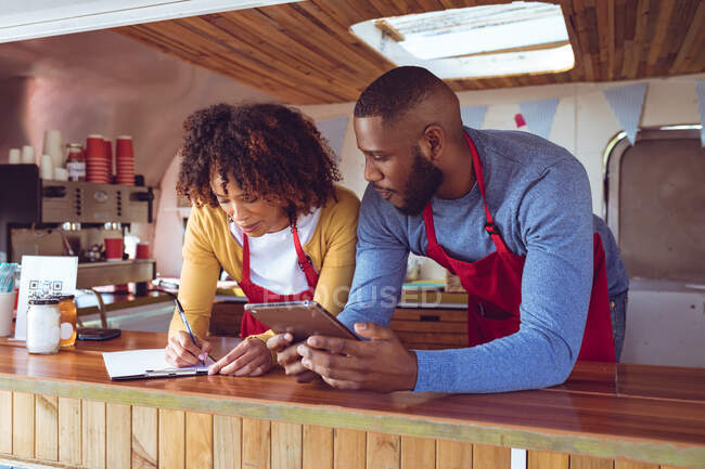 Smiling diverse couple behind counter using tablet and writing in food truck. independent business and street food service concept. — Stock Photo