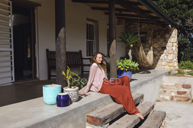 Femme caucasienne souriante assise sur des marches se relaxant sur la terrasse ensoleillée du chalet. mode de vie sain, proche de la nature dans la maison rurale hors réseau. — Photo de stock