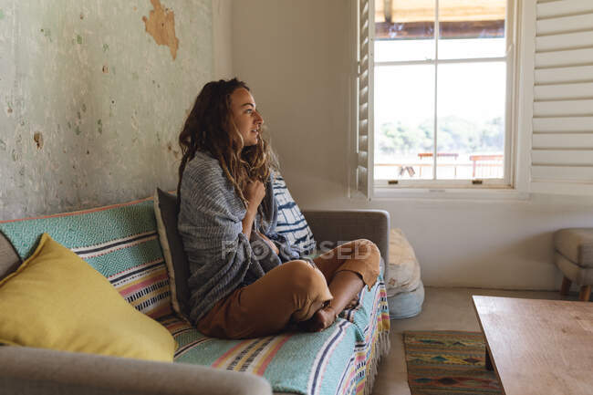 Felice donna caucasica indossando coperta sopra le spalle seduto sul divano in soleggiato soggiorno cottage. semplice vivere in una casa rurale fuori dalla griglia. — Foto stock