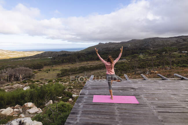 Visão traseira da mulher caucasiana praticando ioga de pé em uma perna no cenário de montanha rural. vida saudável, fora da grade e perto da natureza. — Fotografia de Stock