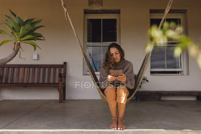 Femme caucasienne assise dans un hamac sur la terrasse du chalet, en utilisant une tablette. mode de vie sain, proche de la nature dans la maison rurale hors réseau. — Photo de stock