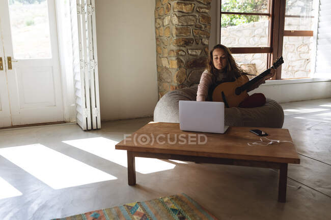Kaukasische Frau sitzt auf Sitzsack und spielt Akustikgitarre mit Laptop im sonnigen Hüttenwohnzimmer. einfaches Leben in einer netzunabhängigen ländlichen Wohnung. — Stockfoto