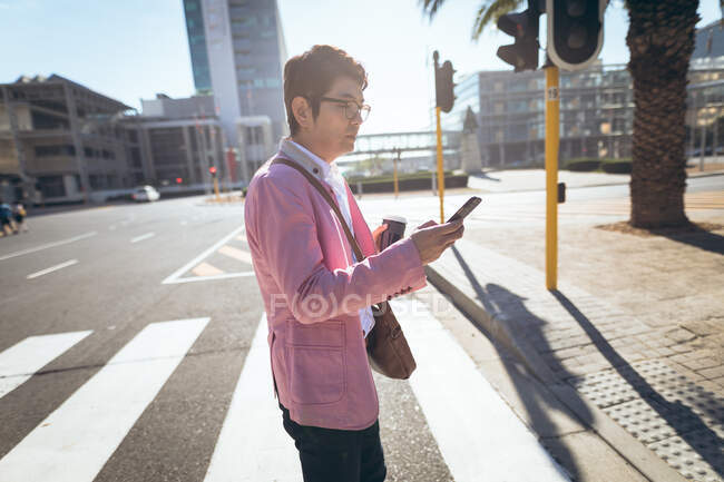 Asian businessman using smartphone drinking takeaway coffee crossing city street. digital nomad out and about in city concept. — Stock Photo