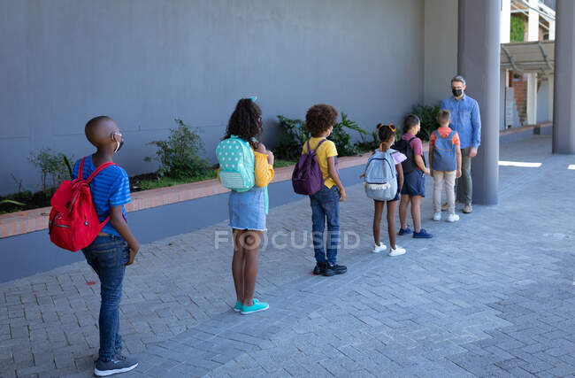 Caucasian male teacher measuring temperature of group of students standing in a queue at school. hygiene and social distancing at school during covid 19 pandemic — Stock Photo