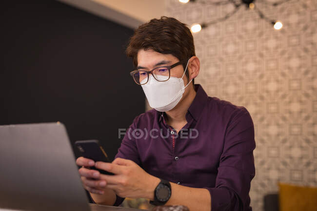 Asian businessman wearing face mask using smartphone and laptop in hotel lobby. digital nomad out and about in city during covid 19 pandemic concept. — Stock Photo